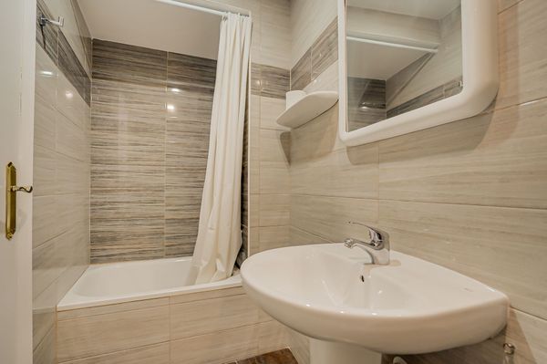 Baño pequeño de color beige con bañera-ducha, lavabo blanco y estantes con espejo en la pared.