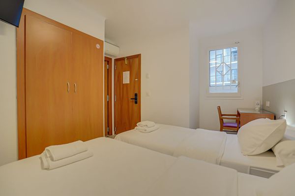 Habitación de hotel pequeña con dos camas individuales, armario de madera, puerta abierta y ventana con una silla.