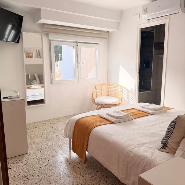 Habitación luminosa con cama, ventana, silla de mimbre, televisor de pared y paredes blancas con detalles en naranja.