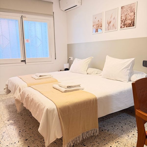 Dormitorio luminoso y minimalista con cama blanca, manta beige, ventana y cuadro enmarcado.