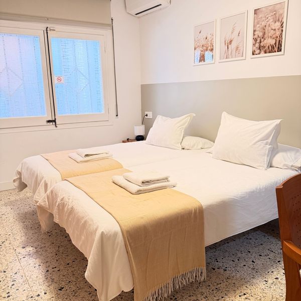 Habitación luminosa con una cama blanca bien hecha, una manta beige, una ventana y cuadros enmarcados en la pared.