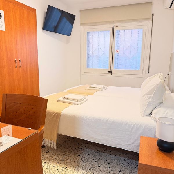 Habitación de hotel pequeña con cama, televisor de pared, ventana, armario y silla de madera.