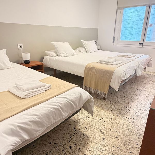 Habitación con dos camas individuales bien hechas, toallas dobladas y una ventana con contraventanas azules.
