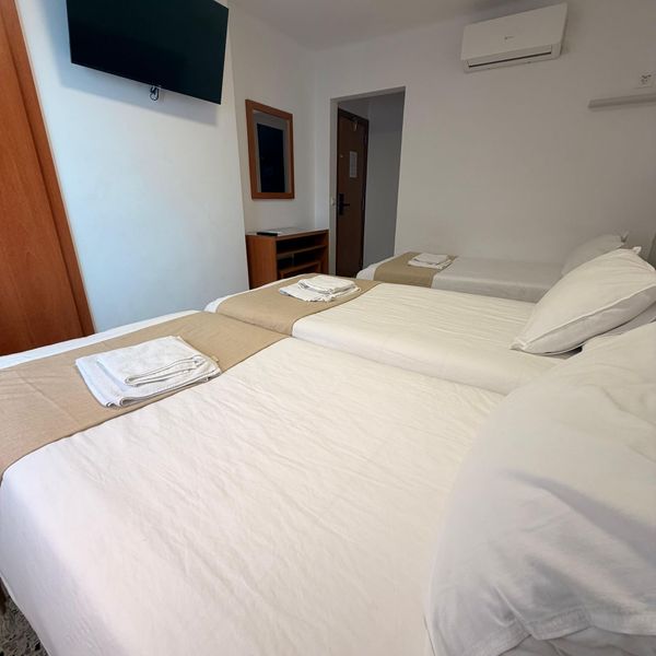 Dos camas individuales en una sencilla habitación de hotel blanca con televisor de pared y aire acondicionado.