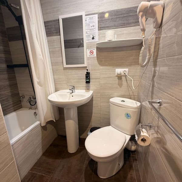 Baño pequeño con bañera, inodoro, lavabo con pedestal, espejo de pared, ducha y paredes de azulejos beige.
