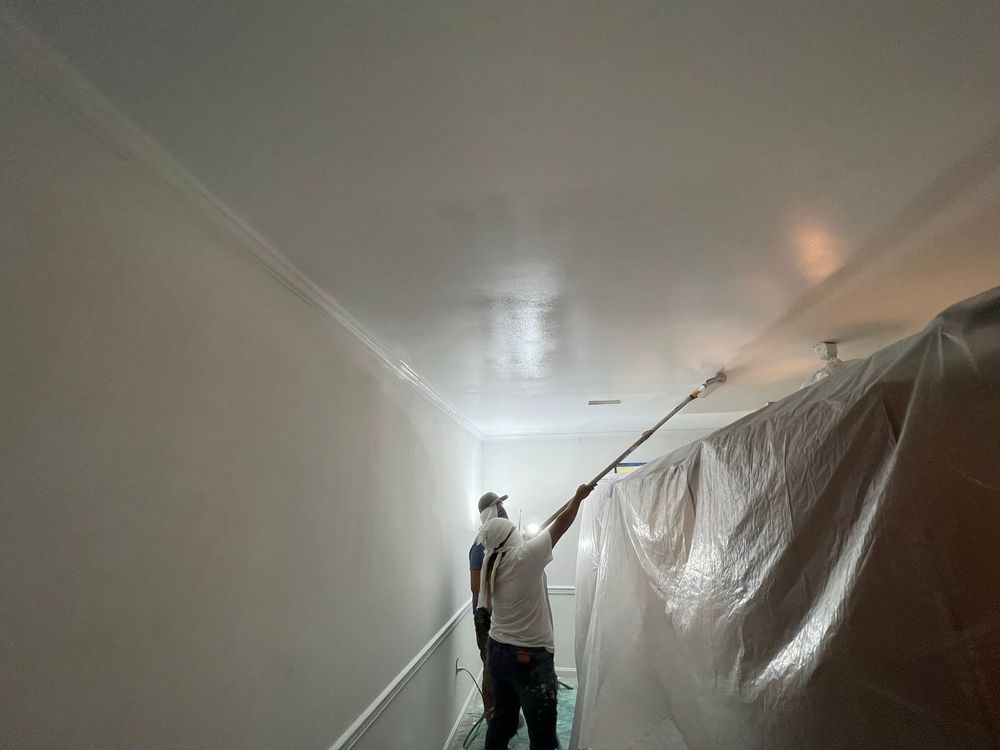 A painter uses an extension pole to spray paint a white ceiling in a room where furniture is covered with plastic sheeting.