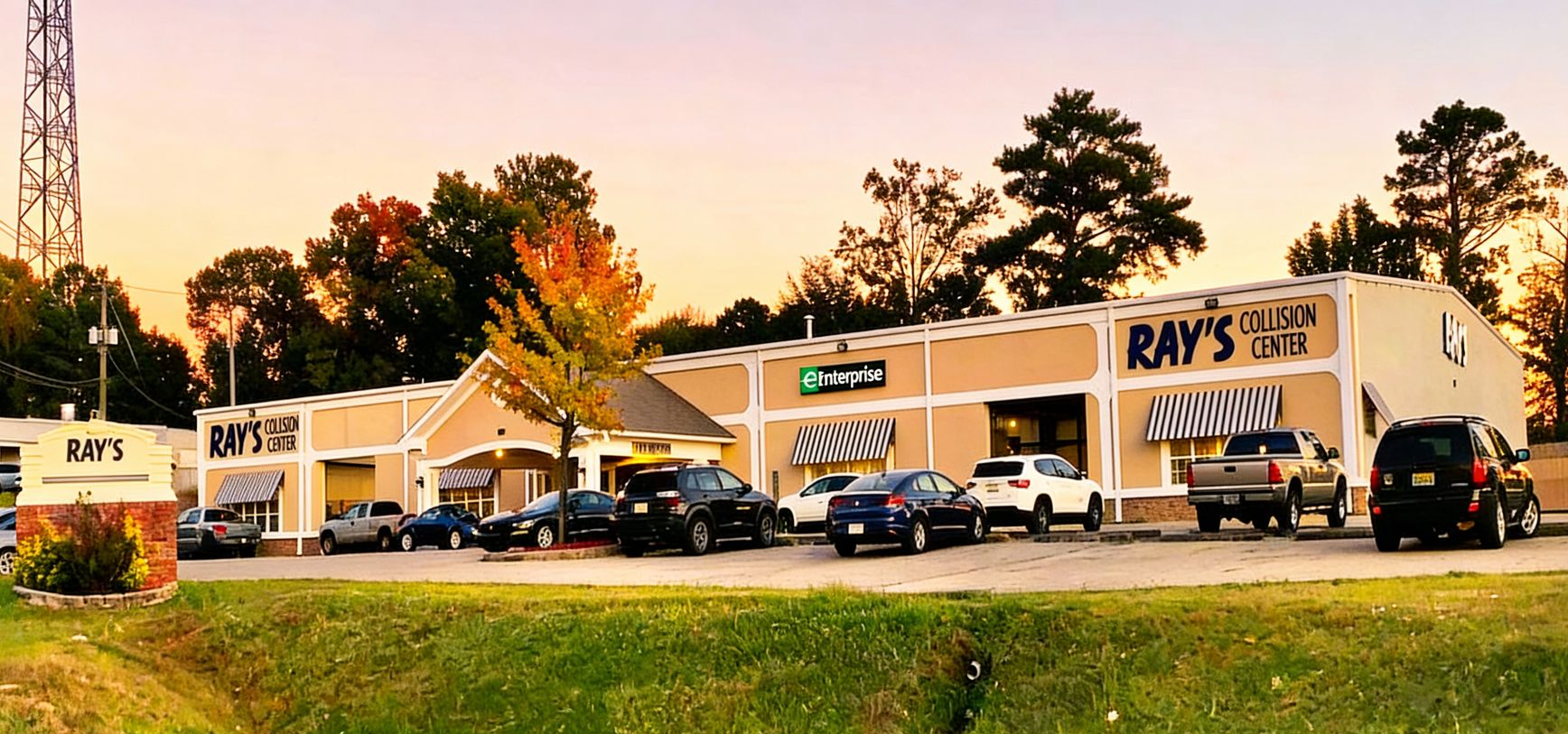 Exterior of a strip mall with a sign for Ray's. Cars parked out front; fall foliage and sunset.