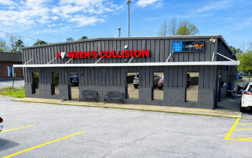 Exterior of a collision repair shop; gray building with red lettering and blue sign; parking lot.