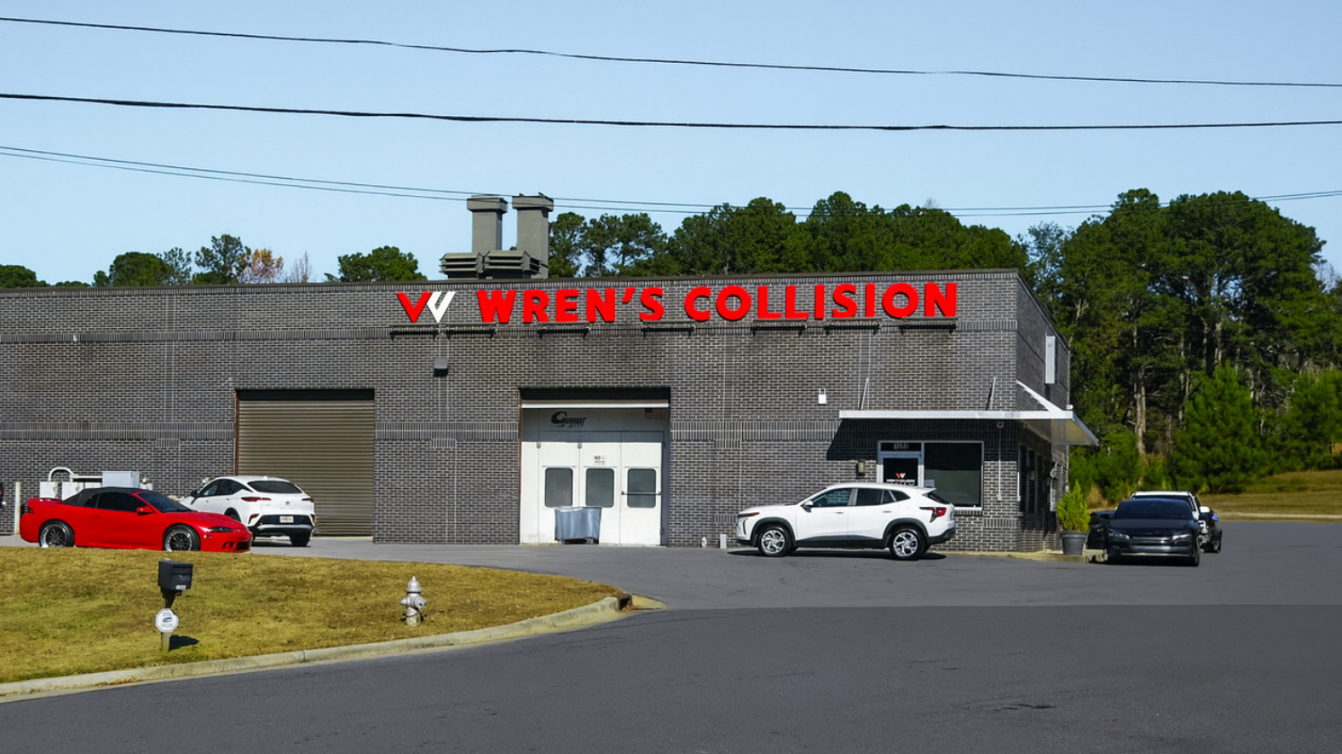Exterior of V. Week's Collision repair shop with cars parked outside. Red sign, gray building, and blue sky.