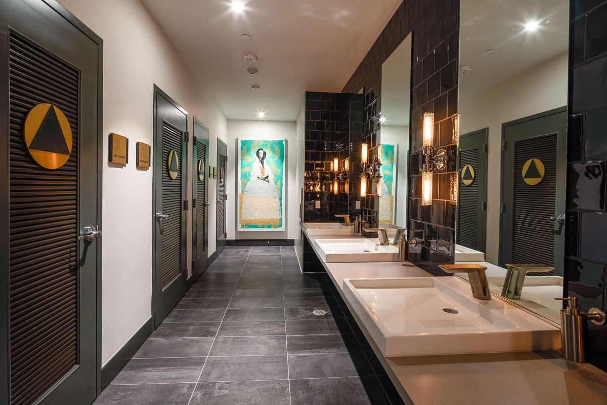 A modern public restroom with sinks, mirrors, and individual stalls. Artwork hangs on the wall, gray tile floor.