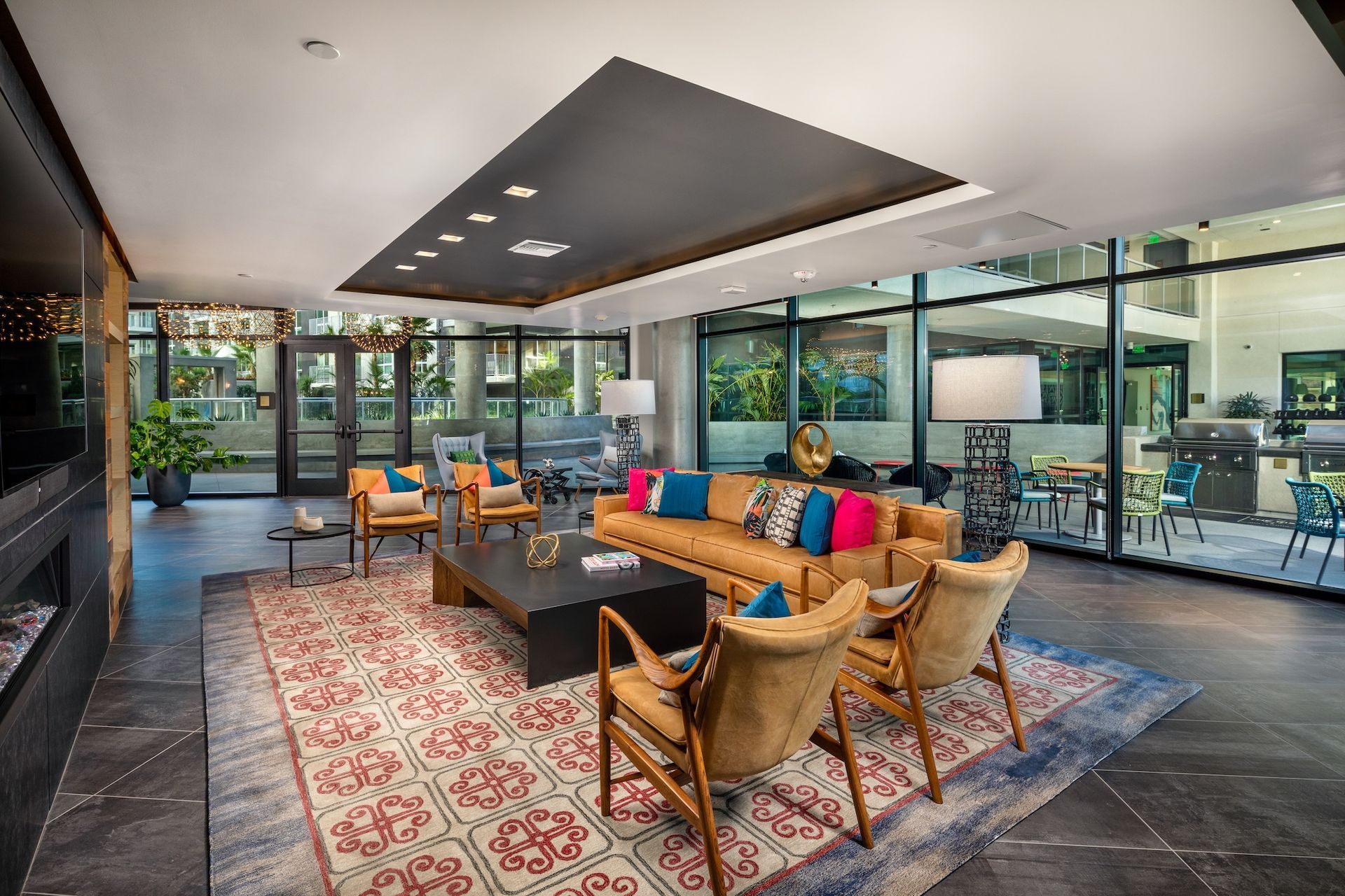 Lounge with a brown leather sofa, armchairs, and a patterned rug. Large windows overlook a patio.