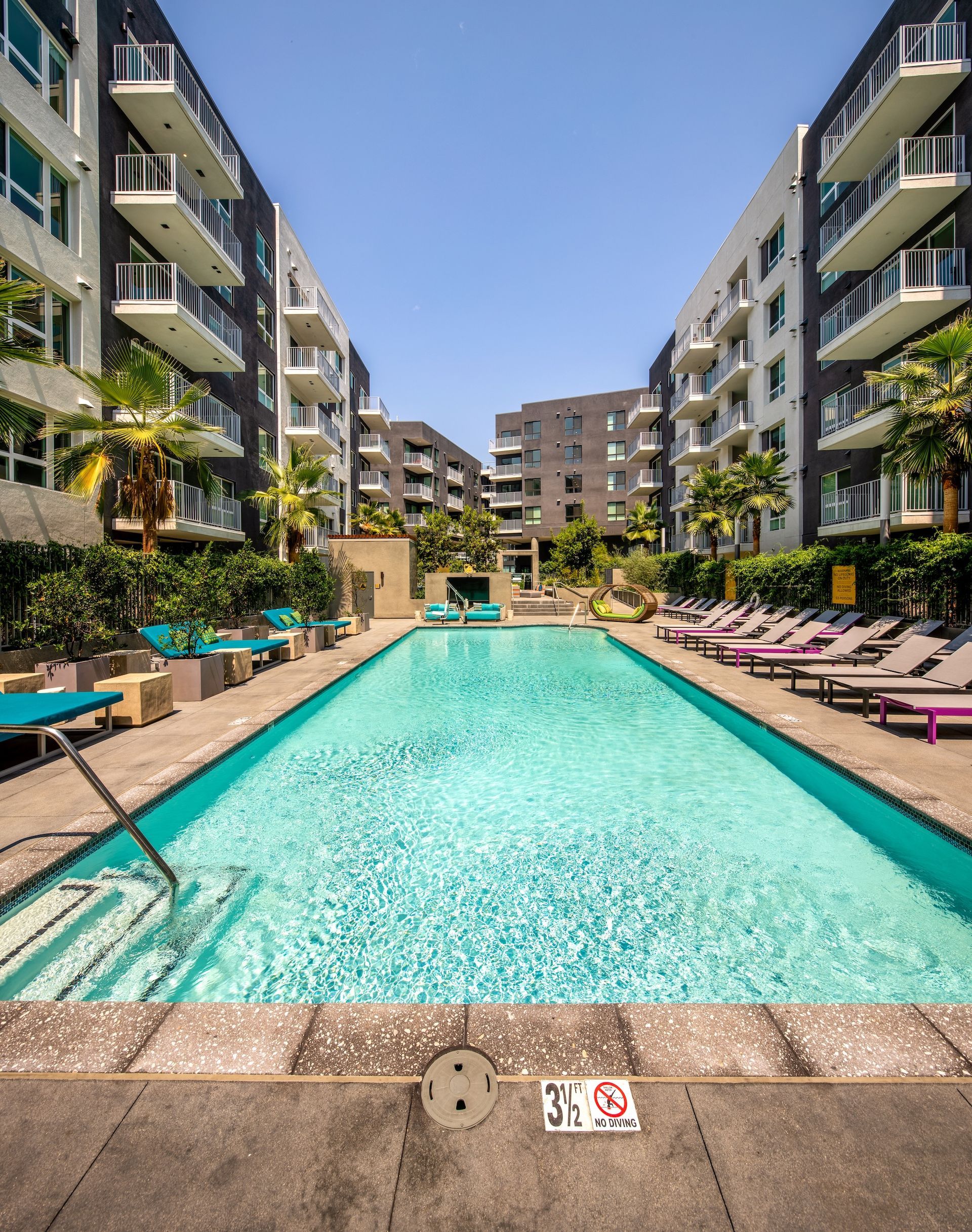 A long, rectangular pool flanked by lounge chairs between two apartment buildings on a sunny day.