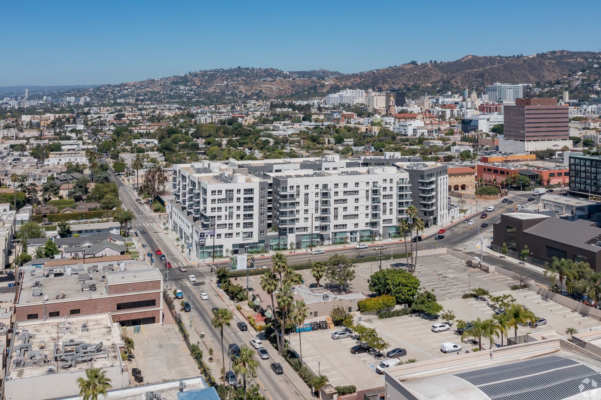 Aerial view of a white apartment building with balconies, palm trees, and city backdrop on a sunny day.