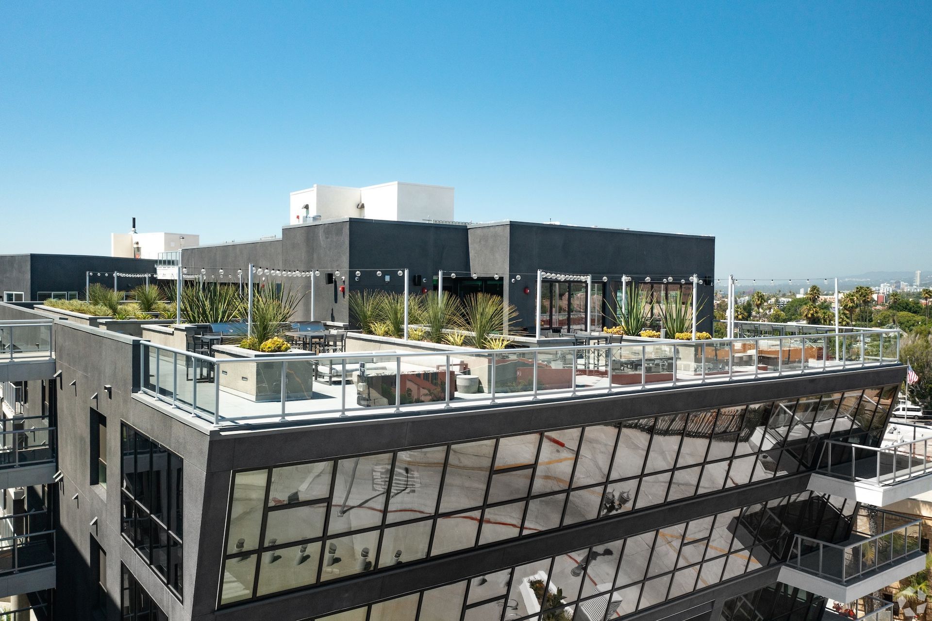 Rooftop terrace on a modern building, featuring glass railings, seating, and landscaping, against a blue sky.