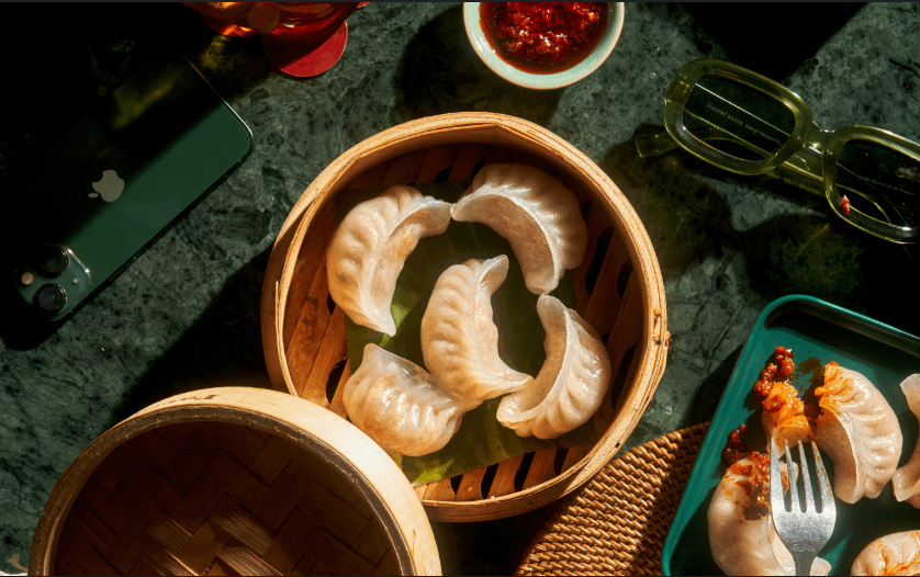 Steamed dumplings in a bamboo basket with dipping sauce, phone, and glasses on a green surface.