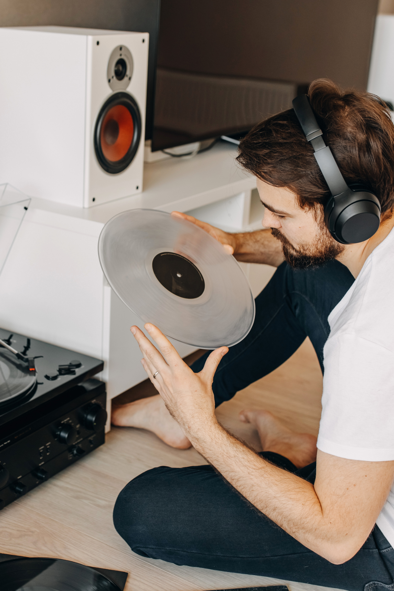 Man inspecting a vinyl record while wearing headphones, near a turntable and speaker.