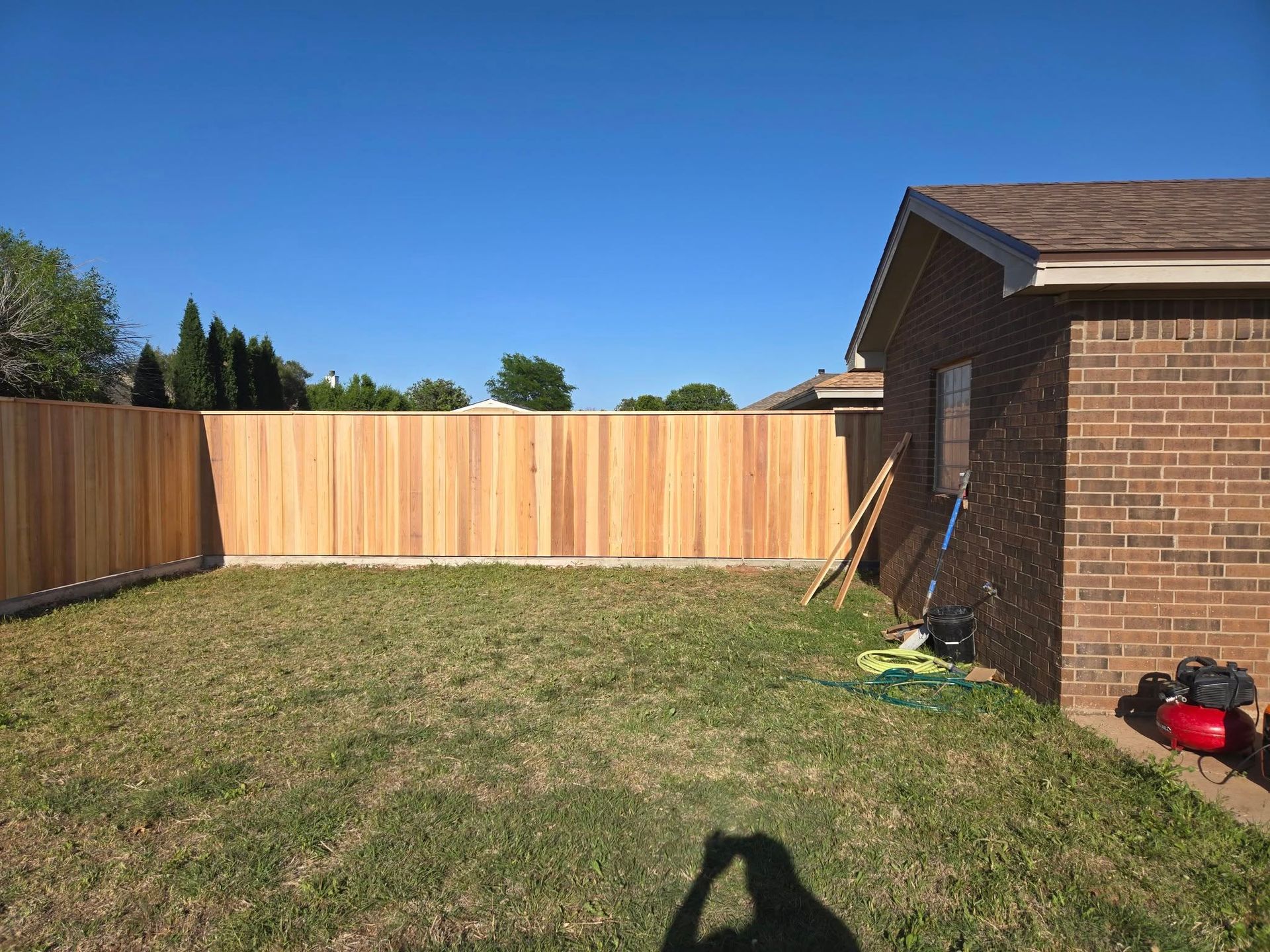 A newly built wooden fence surrounds a backyard with a brick house under a blue sky.