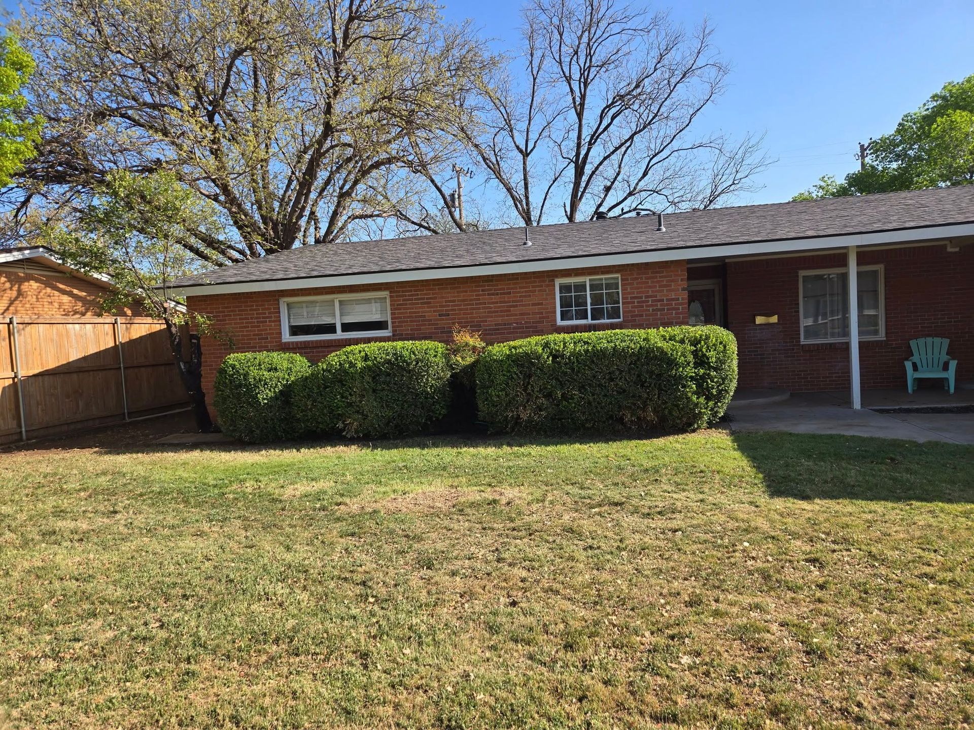 Brick house with trimmed bushes, brown roof, green lawn, and a blue chair.