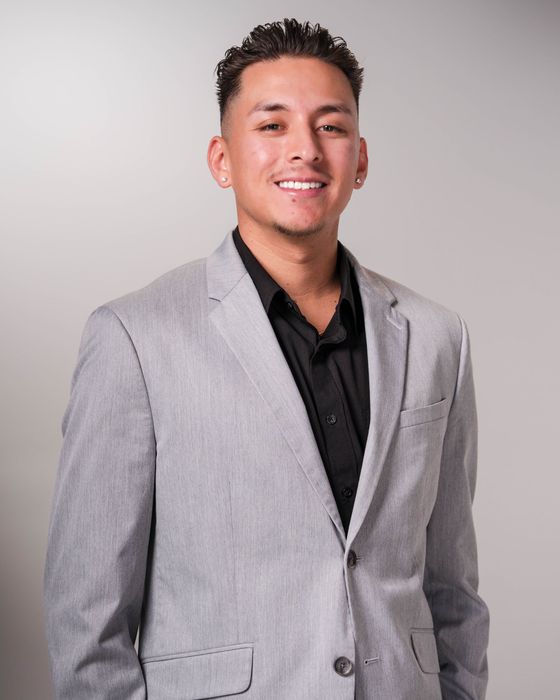 Man in a light gray blazer and black shirt smiling against a plain gray background