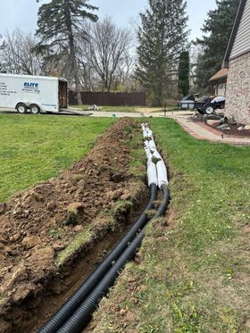 Trenched yard with black drain pipes and white conduit running through grass beside a house.