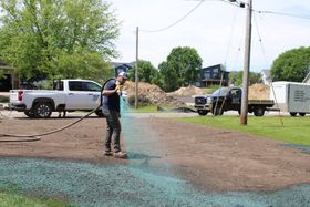 Worker spraying wet surface at a muddy construction site with parked trucks and trees nearby