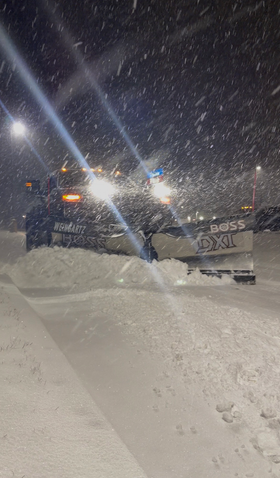 Snowplow clearing a snowy road at night during heavy snowfall, bright lights shining through the snow.