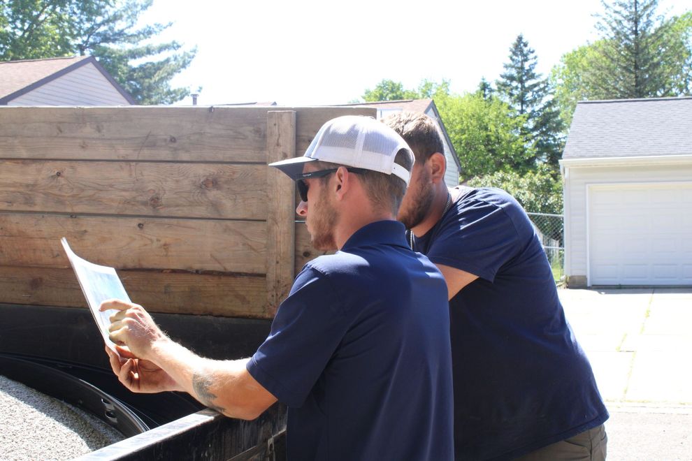 Two workers in navy shirts inspecting paperwork beside a large wooden dumpster outdoors.