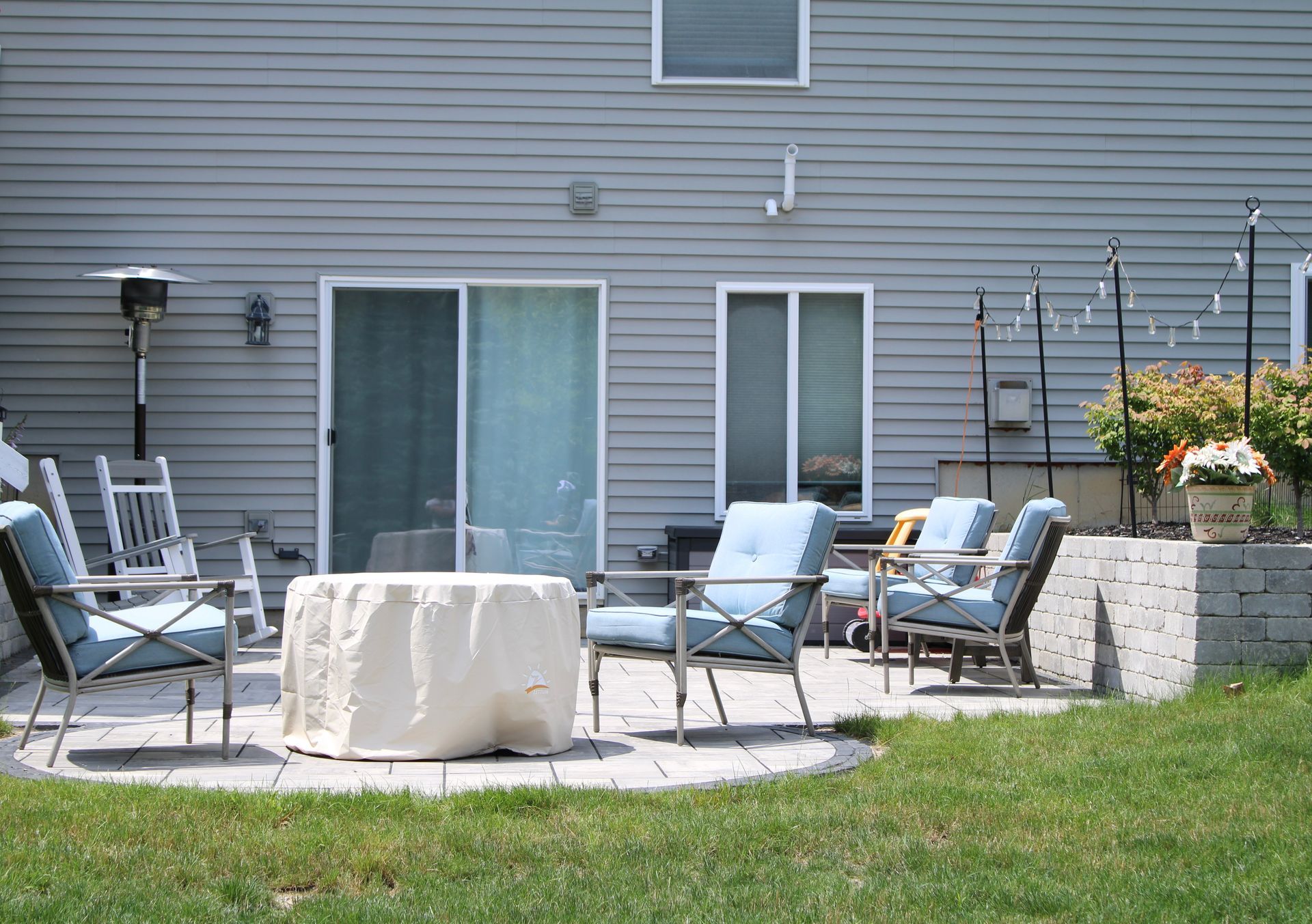 Backyard patio with metal chairs, table, and grill beside a house and flower planters.