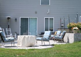 Backyard patio with metal chairs, table, and grill beside a house and flower planters.
