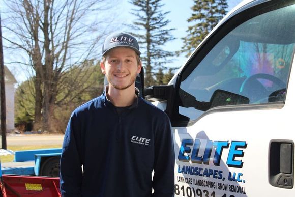Man standing beside a white truck with ELITE Landscaping lettering, outdoors on a sunny day.