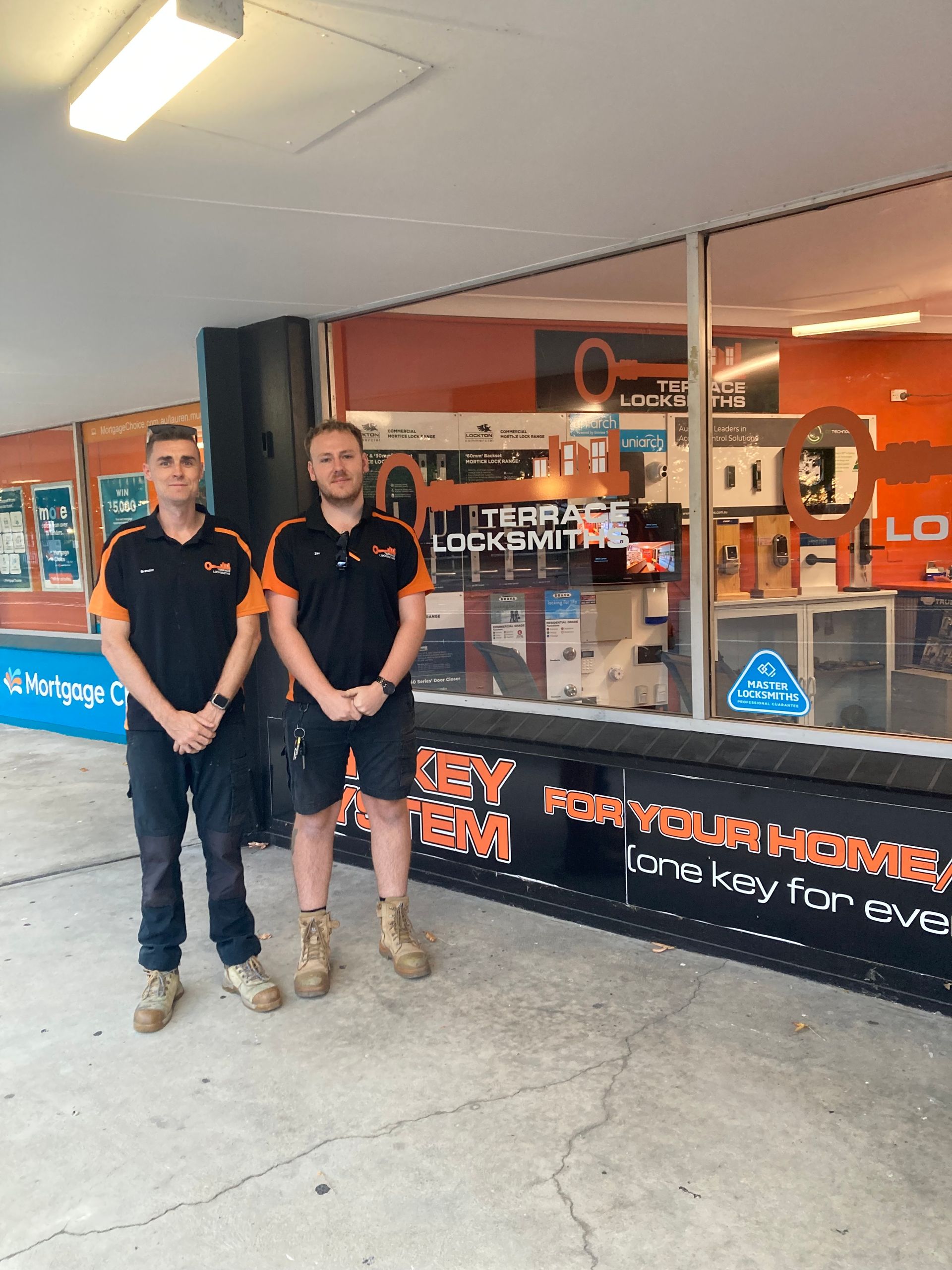 Two staff members in black uniforms standing outside a — Terrace Locksmiths in Raymond Terrace, NSW store with promotional signage