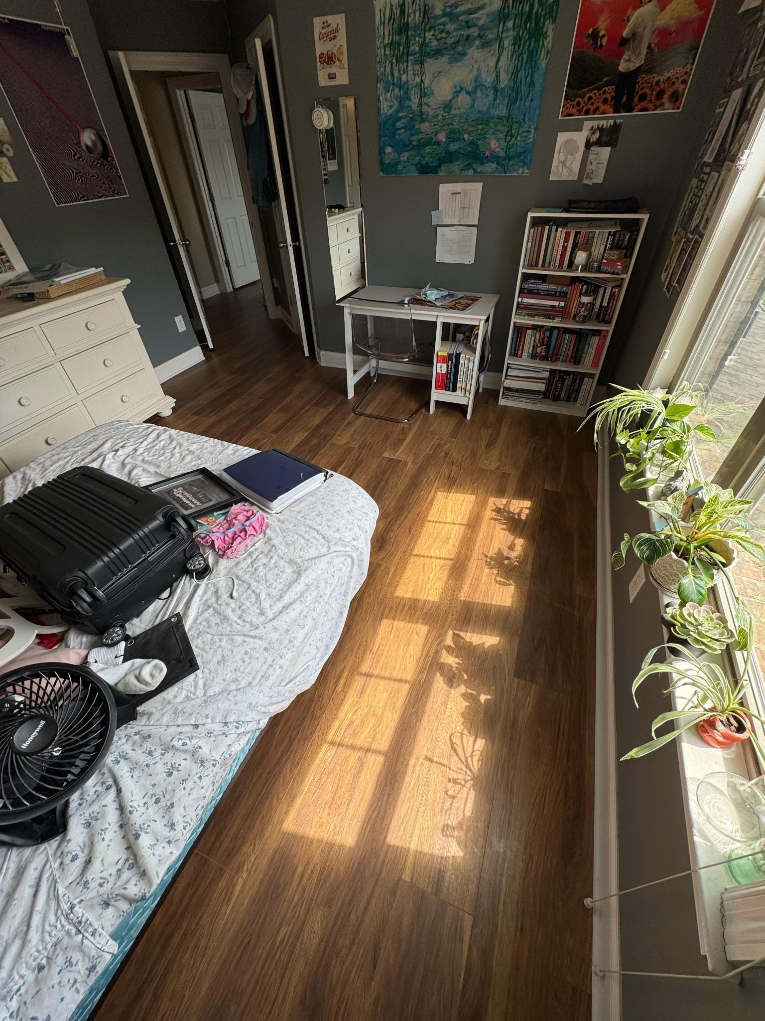 Bedroom with bed, desk, shelving, and window plants; sunlight streams across the wooden floor.
