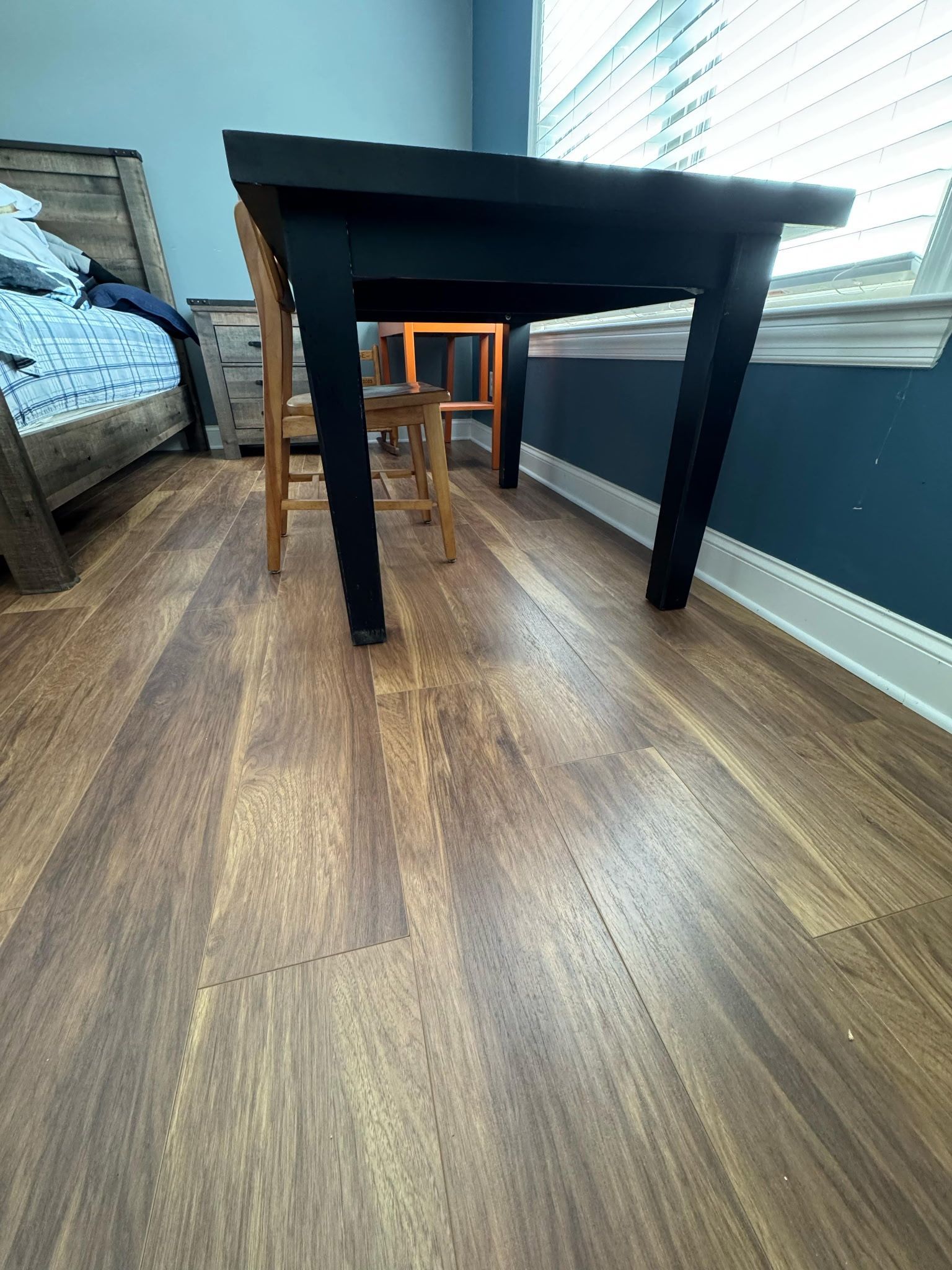 Black table with two wooden chairs on a wooden floor, next to a window with blinds.