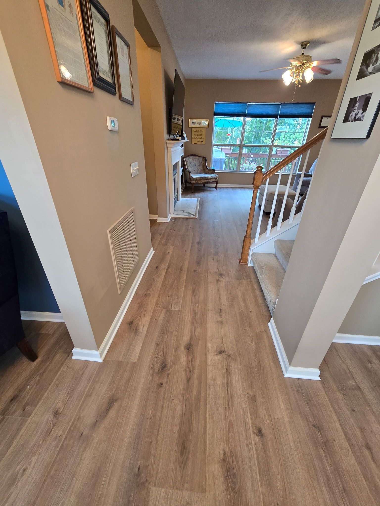 Hallway with light brown flooring, tan walls, and a staircase. Frames and art hang on the walls.