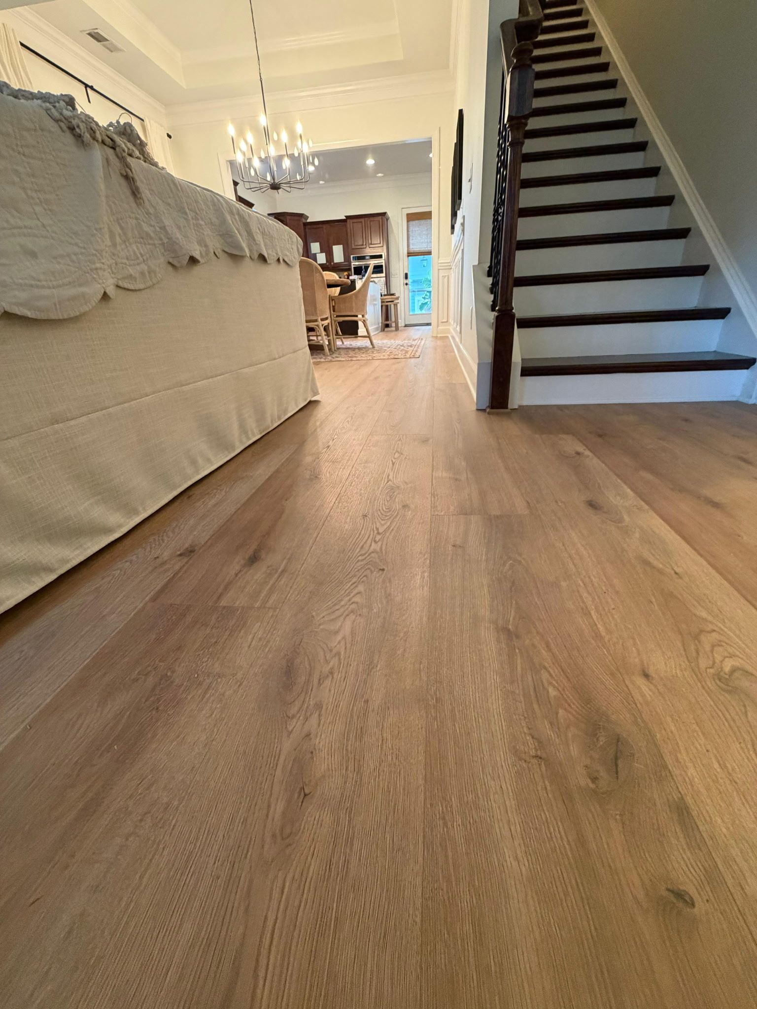 Light-colored hardwood floor in a home, leading towards a staircase and kitchen. Sofa and chandelier visible.