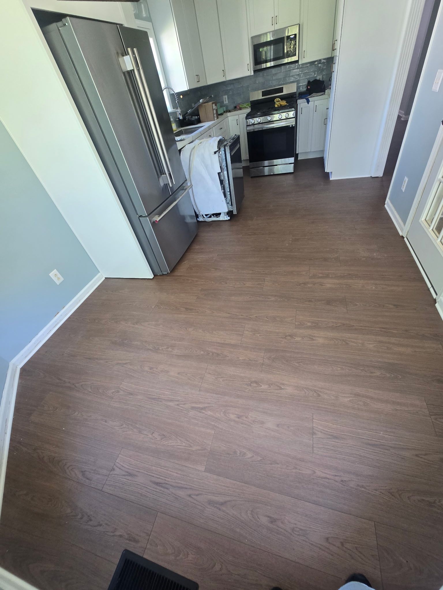 Empty kitchen with stainless steel fridge, white cabinets, and brown wood flooring.