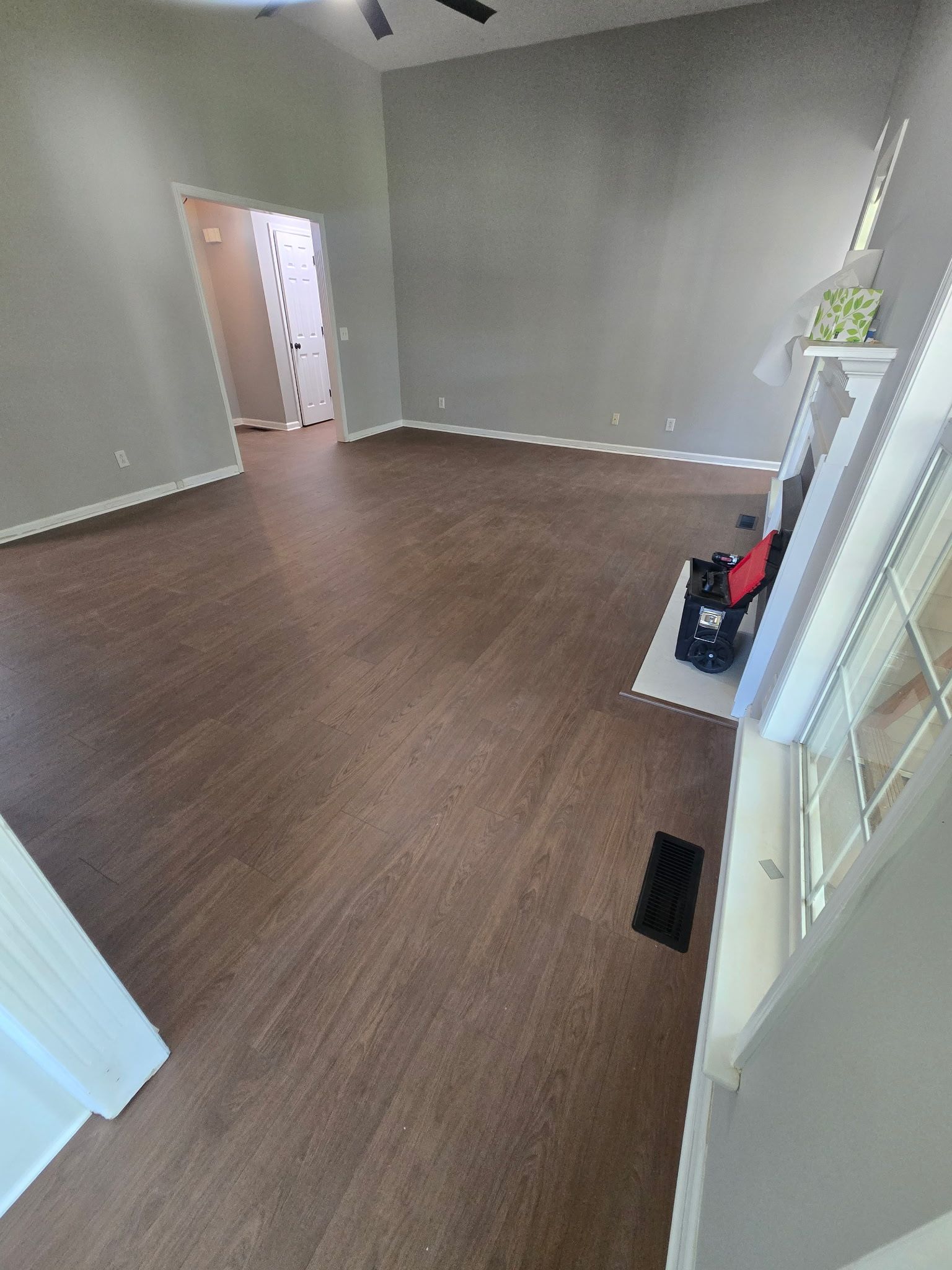 Empty living room with brown wood floors, gray walls, and a doorway to a hall; a red vacuum leans near the window.