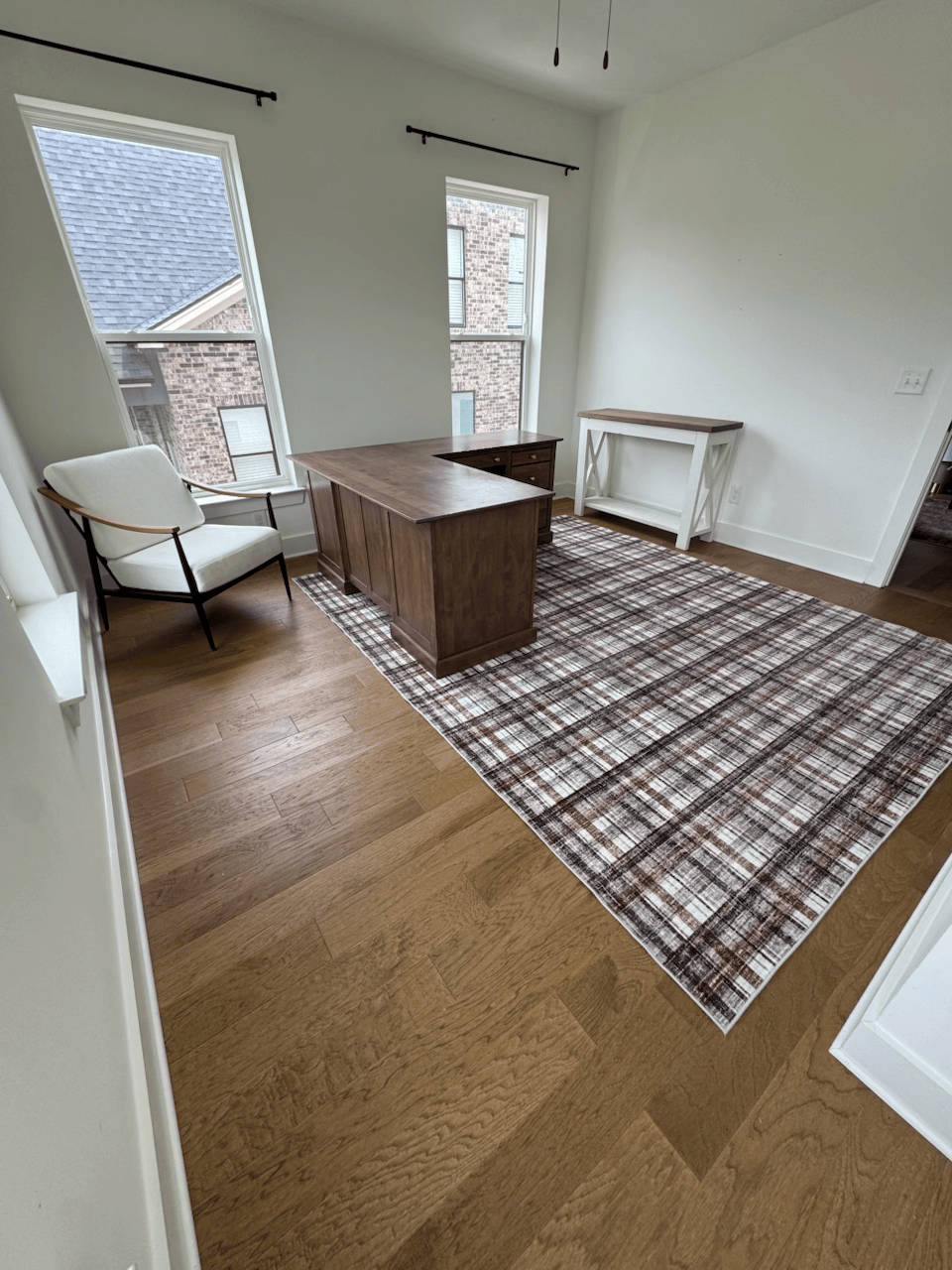 Bright empty room with wood floor, plaid rug, chair, desk, and two windows.