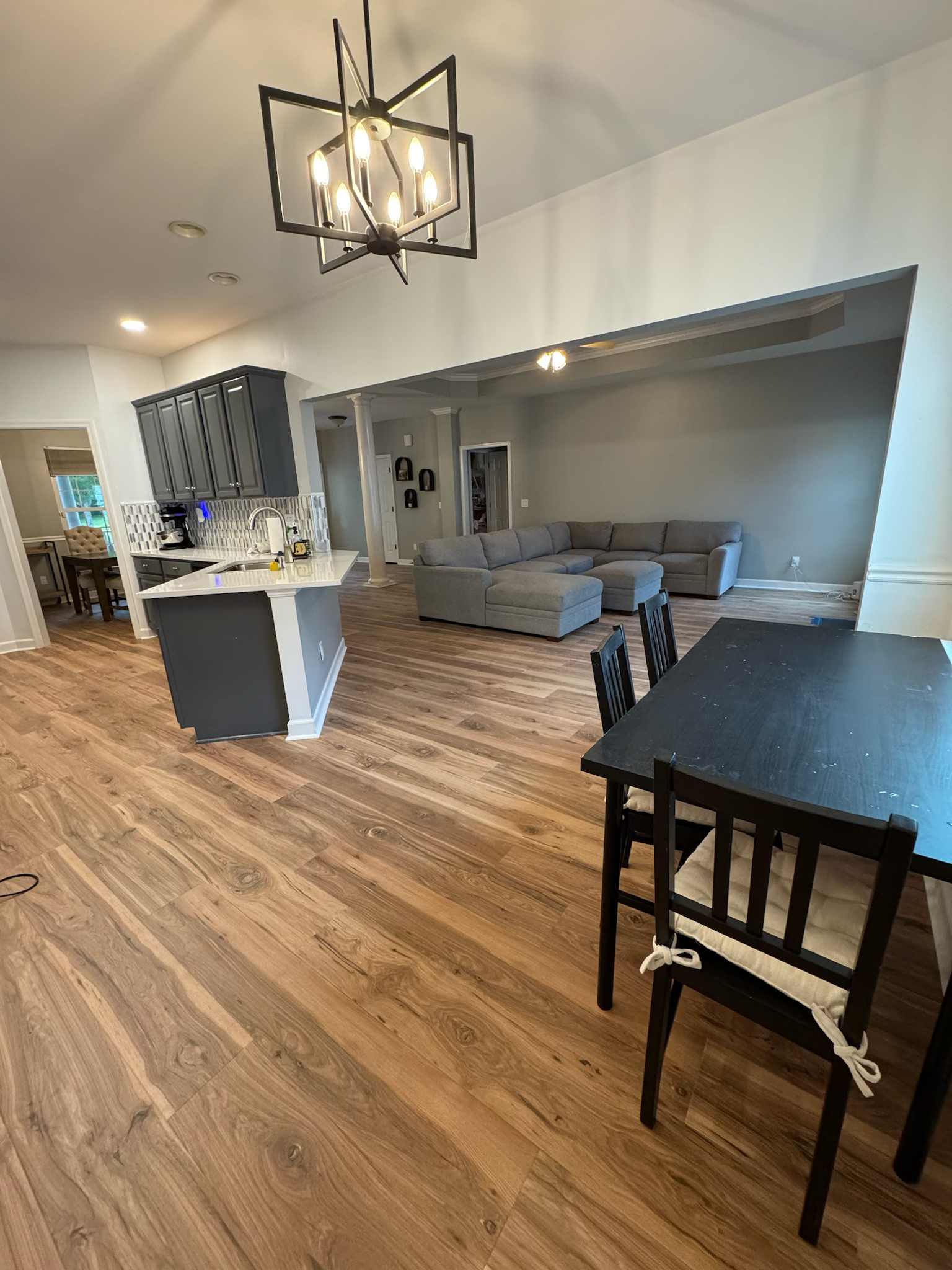 Open kitchen and living room with wood floors, gray sectional sofa, and black dining table under a chandelier.
