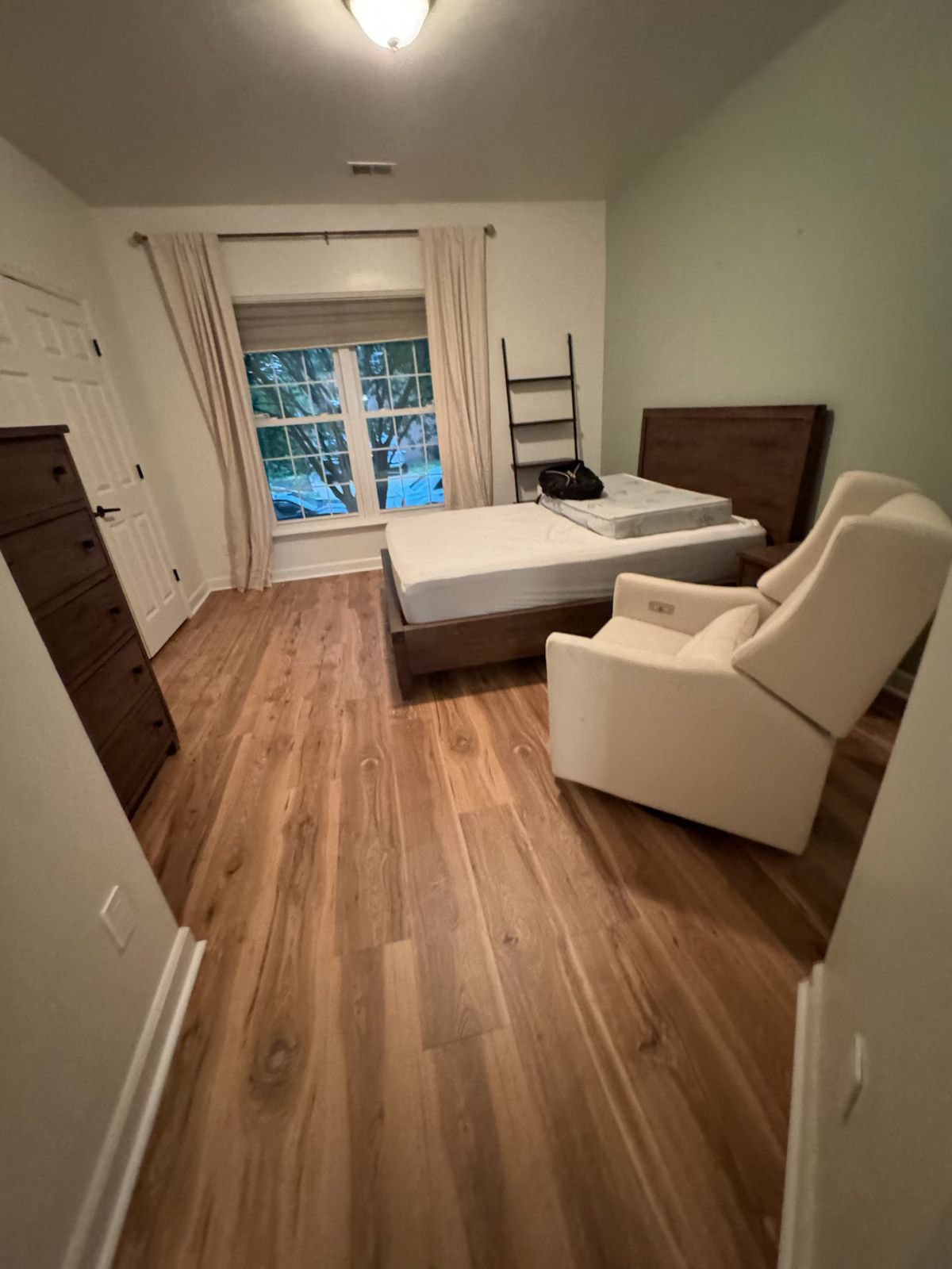 Bedroom with wood floors, white bed and armchair, dresser, and window with curtains