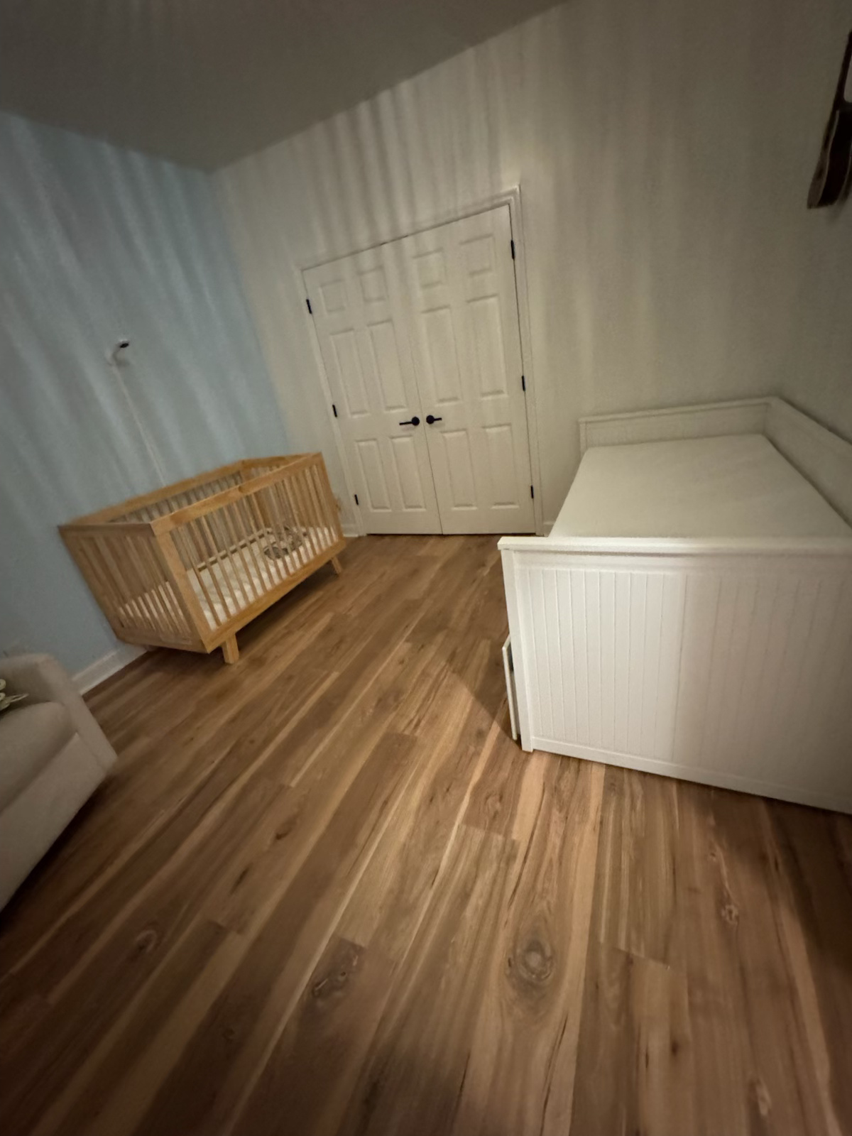 Empty nursery with a white crib, dresser, and chest, wood floor, and closed double doors.