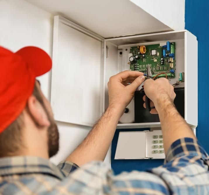 A Man In A Red Hat Is Working On A Security System — FNQ Smoke Alarms & Maintenance In Atherton, QLD