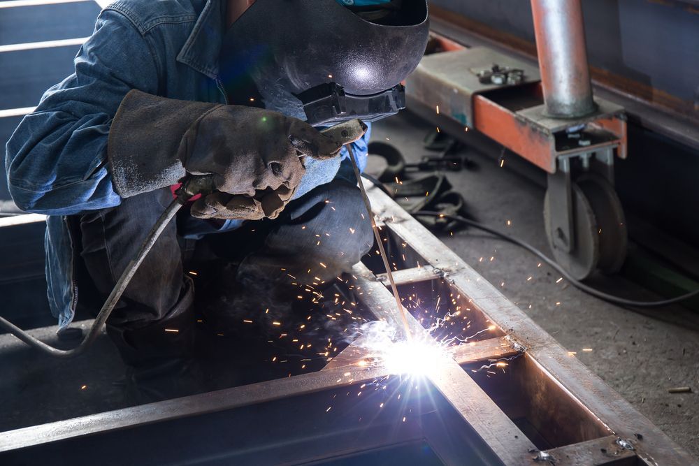 Welder in blue work clothes and a black helmet working on metal frame, sparks flying in a workshop.