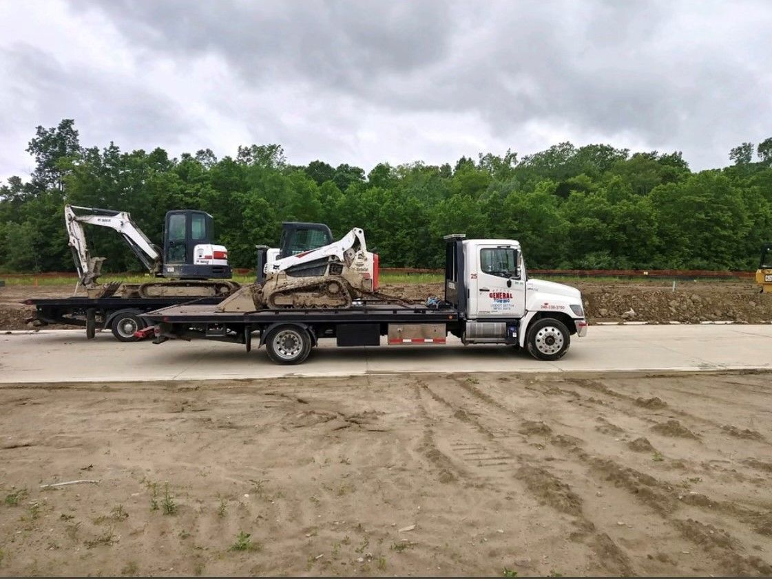 A truck hauling two small excavators on a flatbed trailer on a construction site, cloudy sky background.