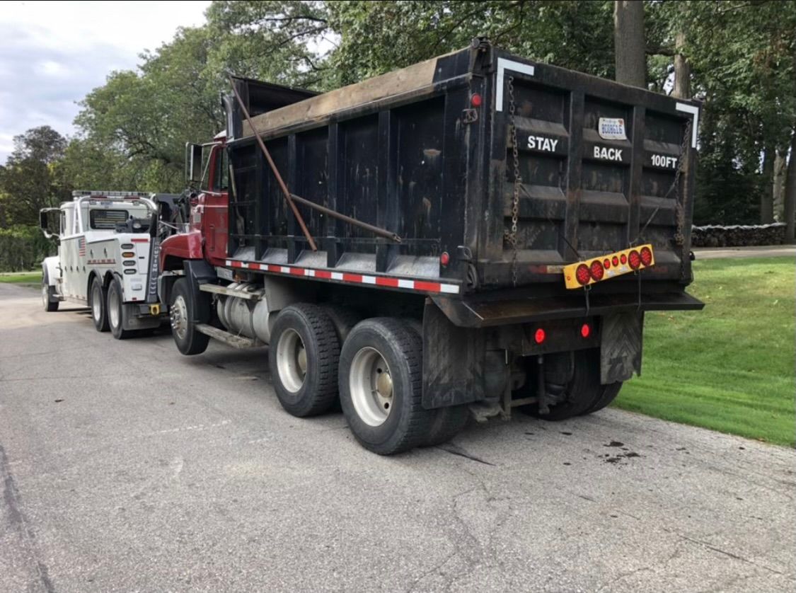 Dump truck on a road, likely transporting materials. Dark brown truck bed, red cab. Bright red and white lights on truck.