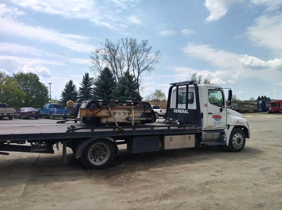 Tow truck with a vehicle part on its flatbed, parked outdoors on a gravel lot.