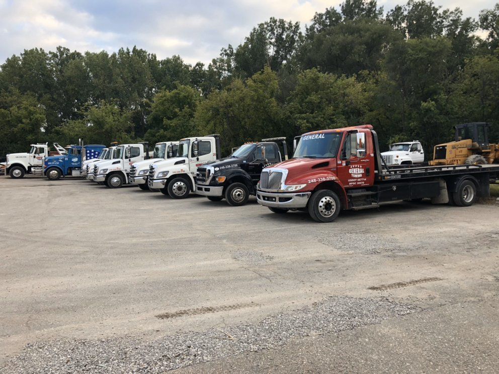 Row of tow trucks parked on a gravel lot in front of trees. Red truck in front.