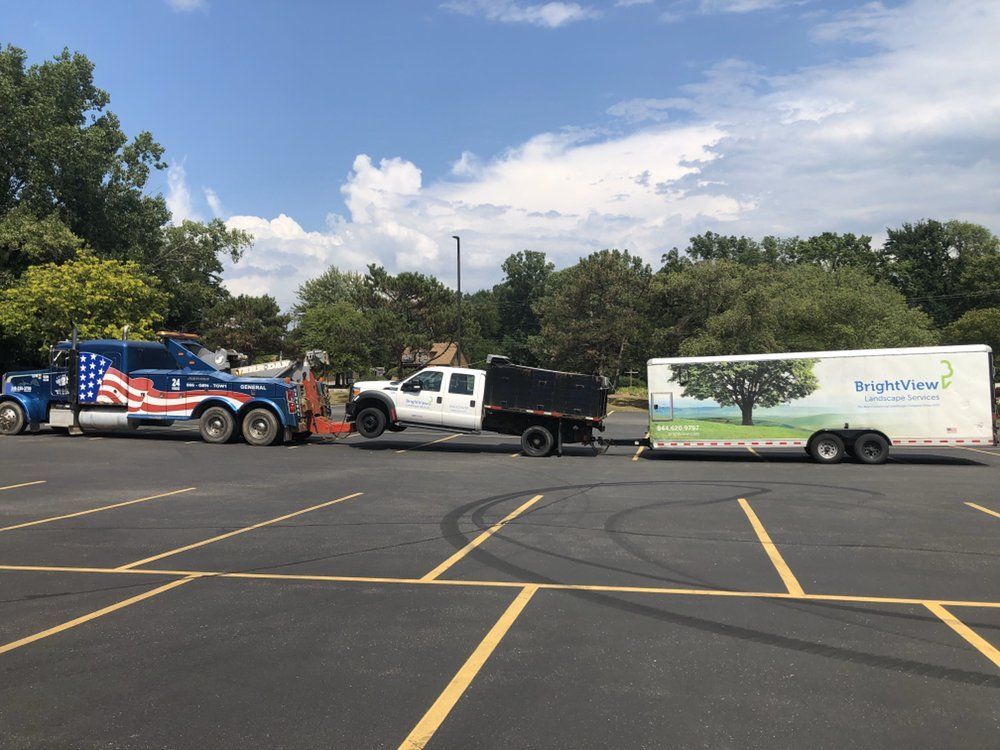 Tow truck towing a pickup truck with trailer in a parking lot on a sunny day.