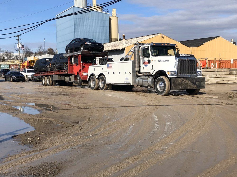 Tow truck hauling multiple cars on a muddy industrial lot, with a blue building and yellow warehouse.