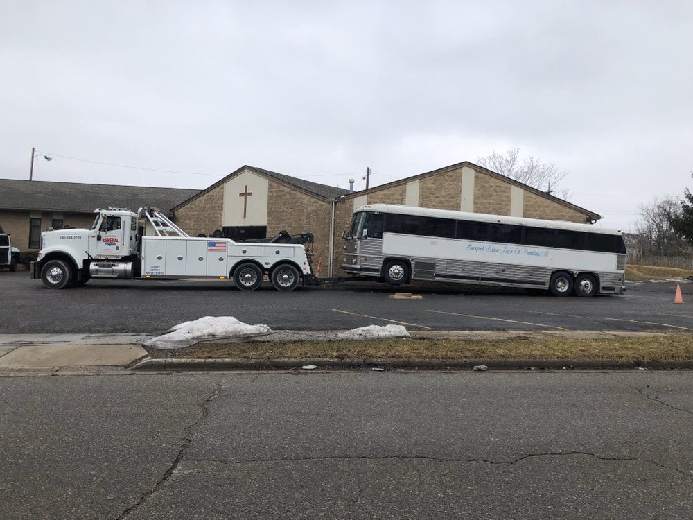 A tow truck pulling a large bus near a brick building with a cross; overcast sky.